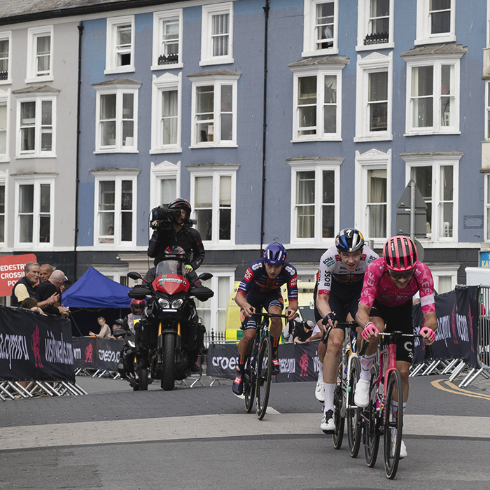 British National Road Championships 2025 - Road Race - Men - a group of riders pass houses painted in differing shades of blue on the seafront in Aberystwyth