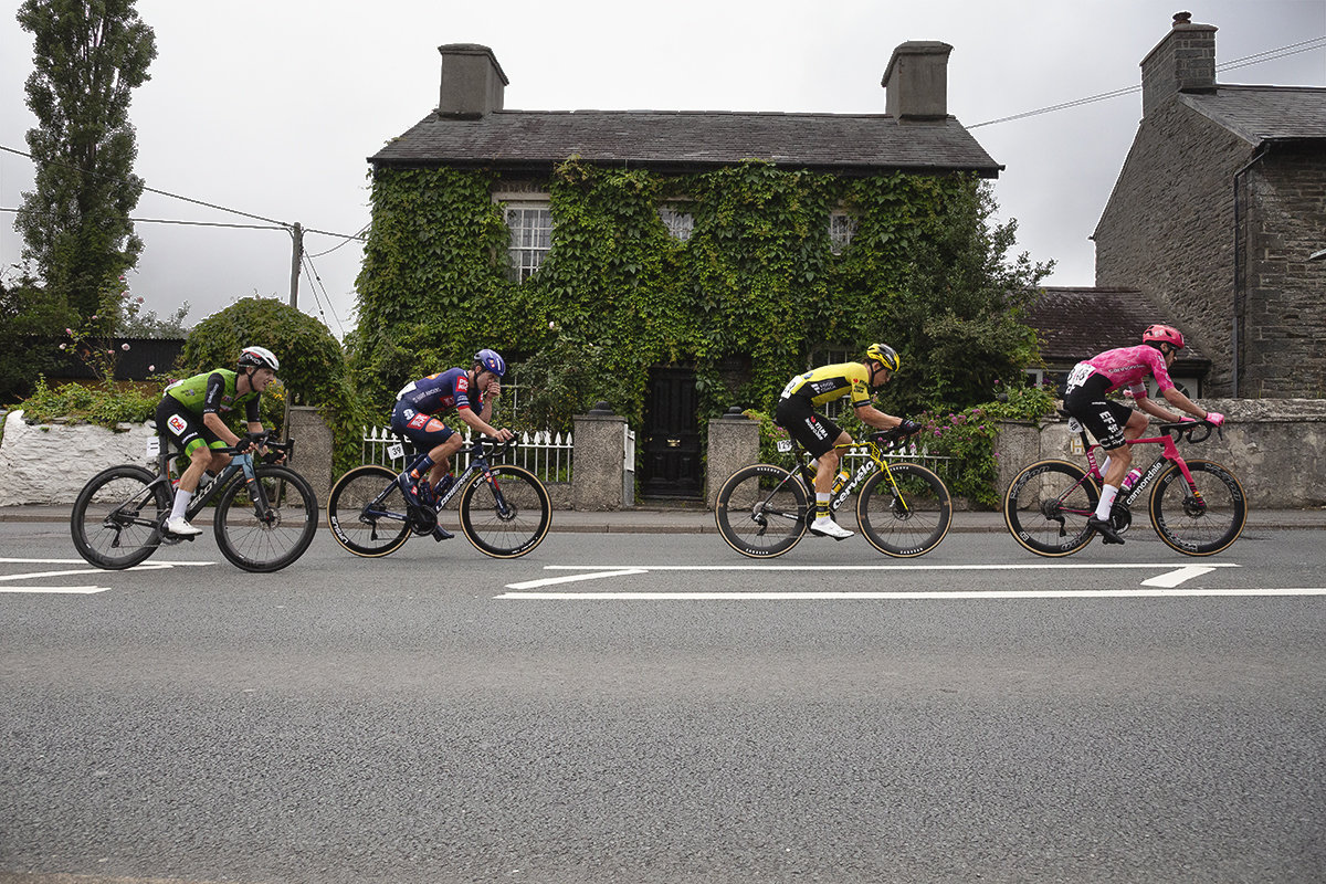British National Road Championships 2025 - Road Race - Men - a group of riders race past a stone cottage covered in ivy in Llanfarian