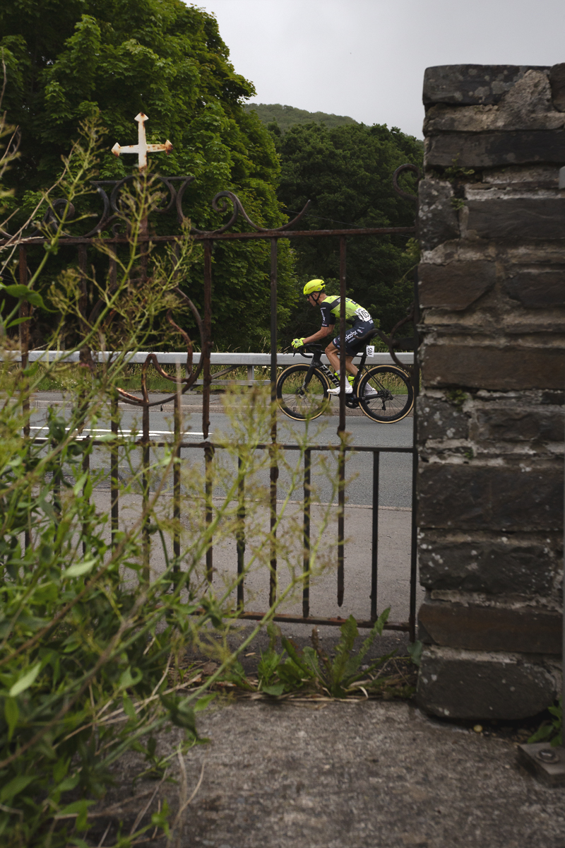 British National Road Championships 2025 - Road Race - Men - Harrison Wood of Sabgal/Anicolor seen through an iron gate with a cross on its top