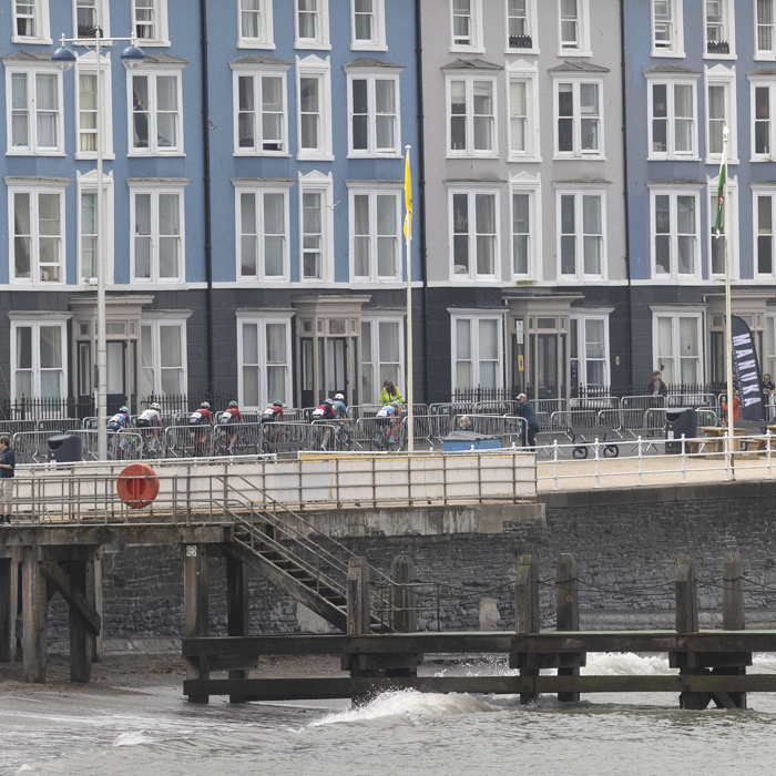 British National Road Championships 2025 - Road Race - Women - Aberystwyth - a group of riders rides past buildings painted in differing shades of blue on the seafront with waves crashing in the foreground