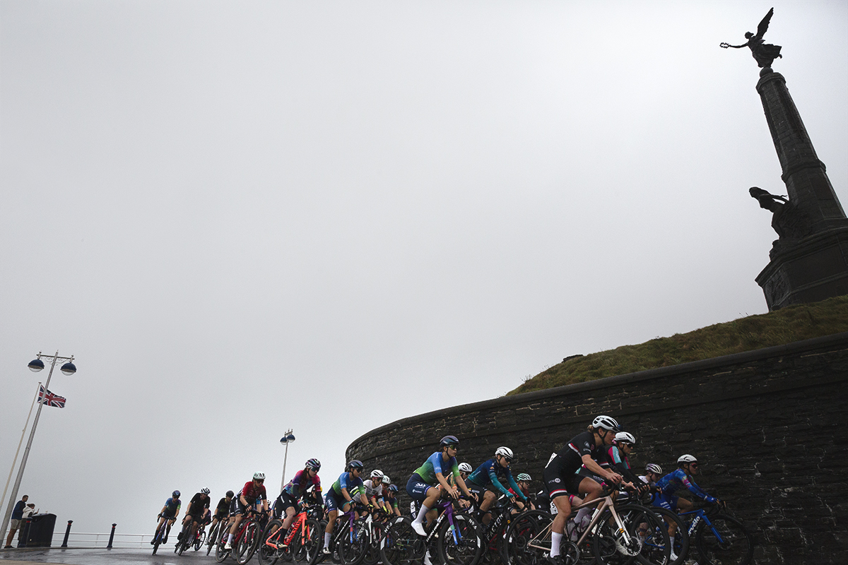 British National Road Championships 2025 - Road Race - Women - Aberystwyth - the peloton rolls out past an obelisk with statues of an angel and woman looking out to sea