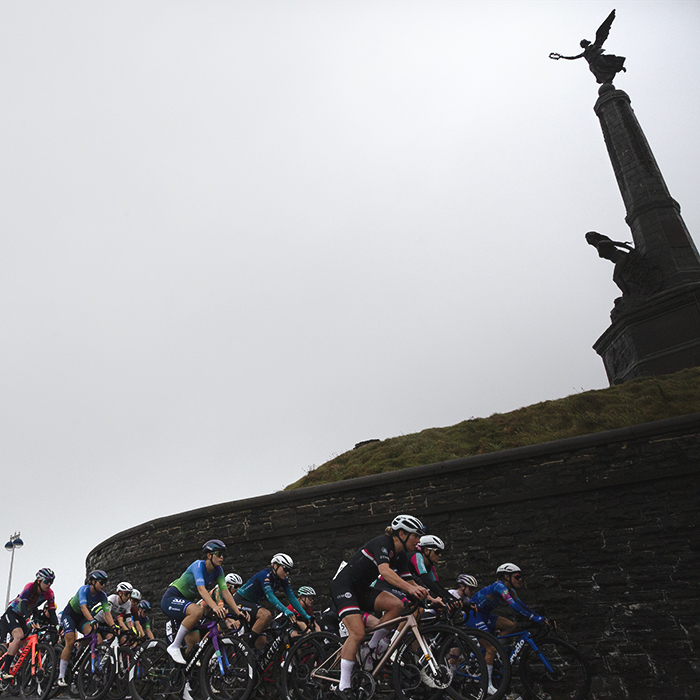 British National Road Championships 2025 - Road Race - Women - Aberystwyth - the peloton rolls out past an obelisk with statues of an angel and woman looking out to sea