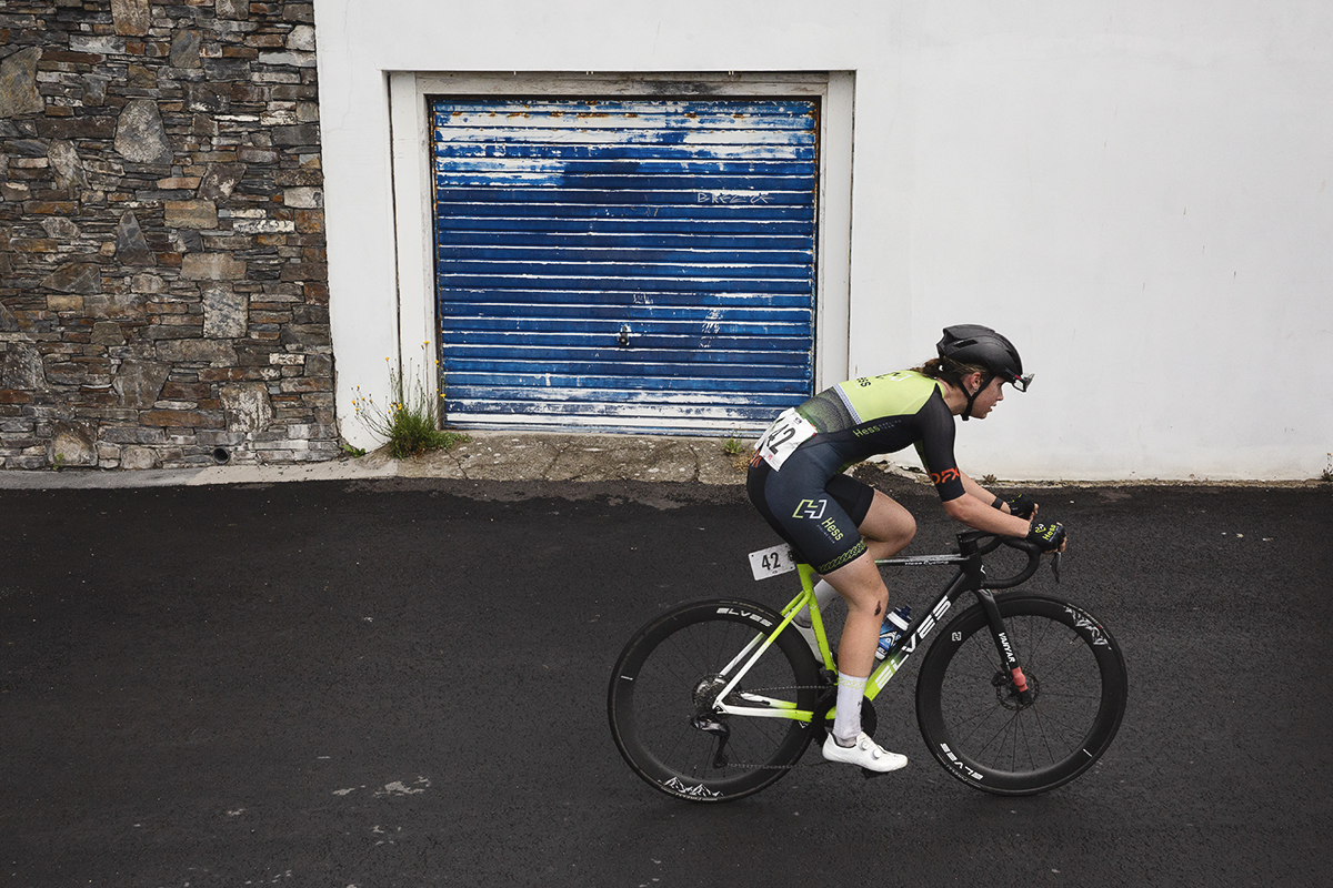 British National Road Championships 2025 - Road Race - Women - Holly Ramsey of Hess Cycling Team rides pass a tired blue garage door on a bright white garage