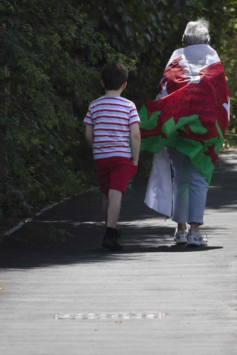 British National Road Championships 2025 - Time Trial - U23 Women - an older women wearing a Welsh flag as a cloak walks along the path accompanied by a young boy