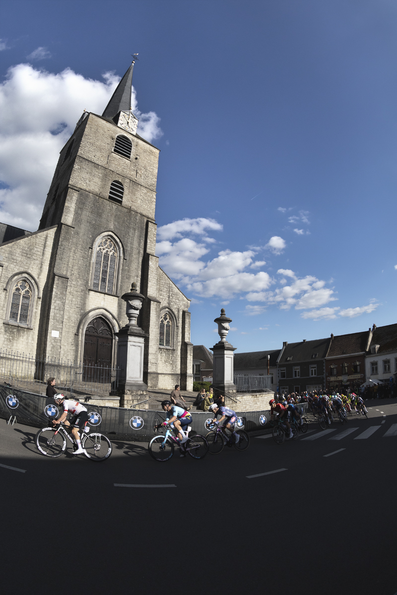 De Brabantse Pijl 2024 - A group of riders emerge from the shadows past Sint-Martinus Kerk and round a corner