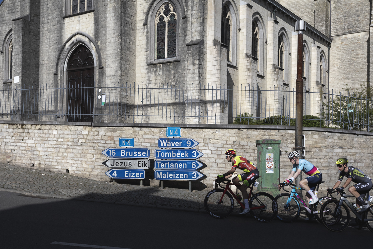 De Brabantse Pijl 2024 - Riders pass road signs to Jezus-Eik outside the church in Overijse