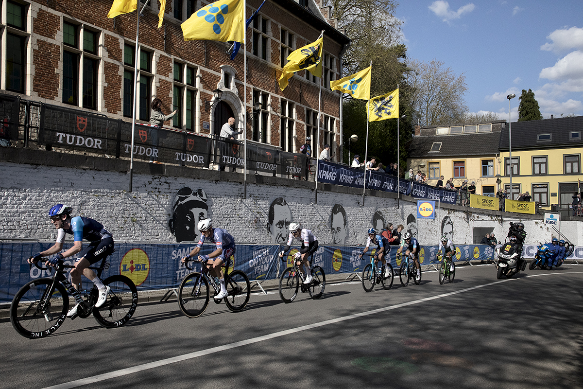 De Brabantse Pijl 2024 - Riders take on the S-Bocht Overijse past a white wall covered in the portraits of past cycling legends