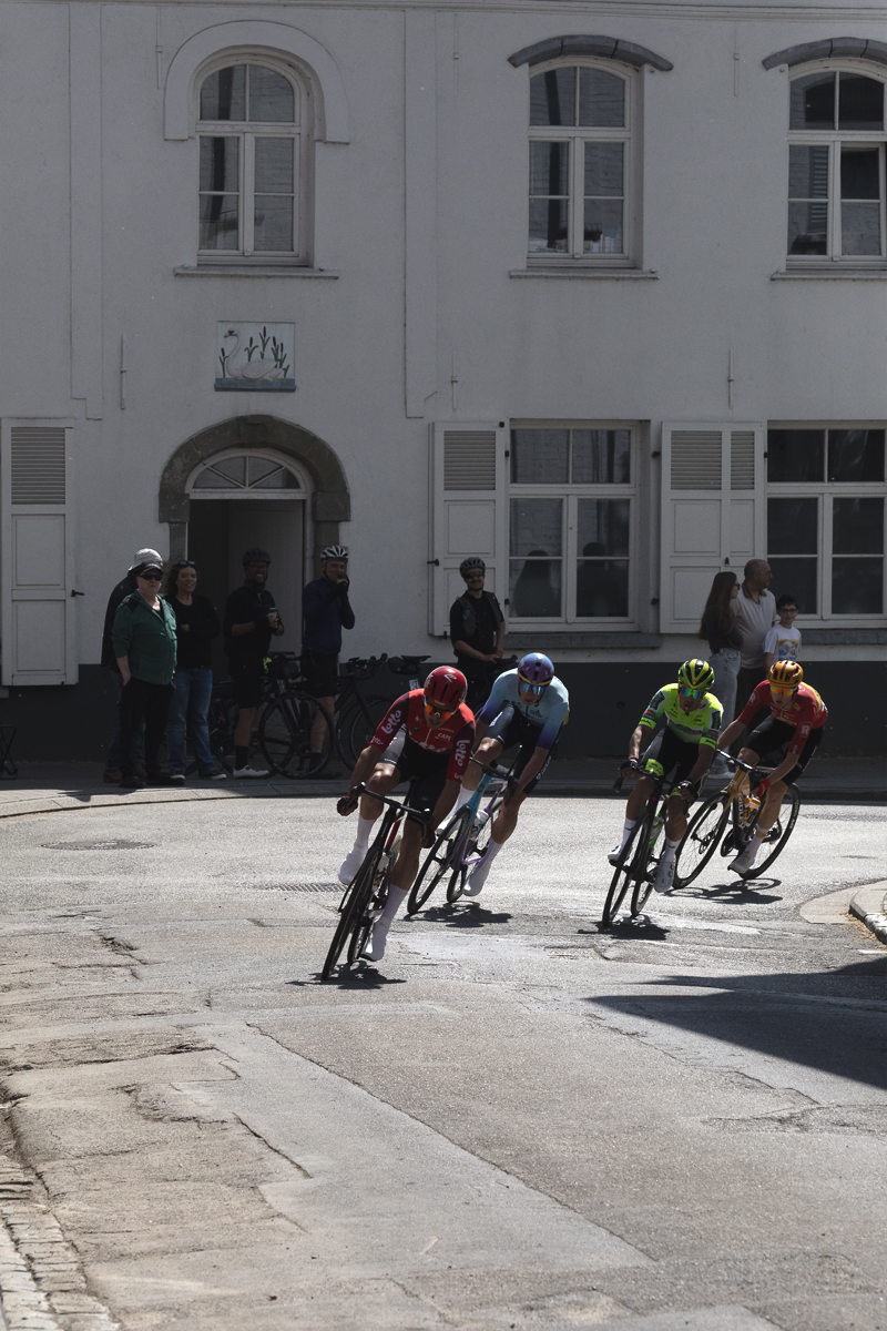 De Brabantse Pijl 2025 - Riders round a corner in Alsemberg with whitewashed buildings and fans in the background