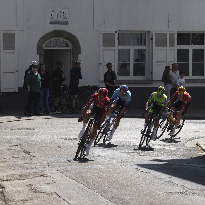 De Brabantse Pijl 2025 - Riders round a corner in Alsemberg with whitewashed buildings and fans in the background