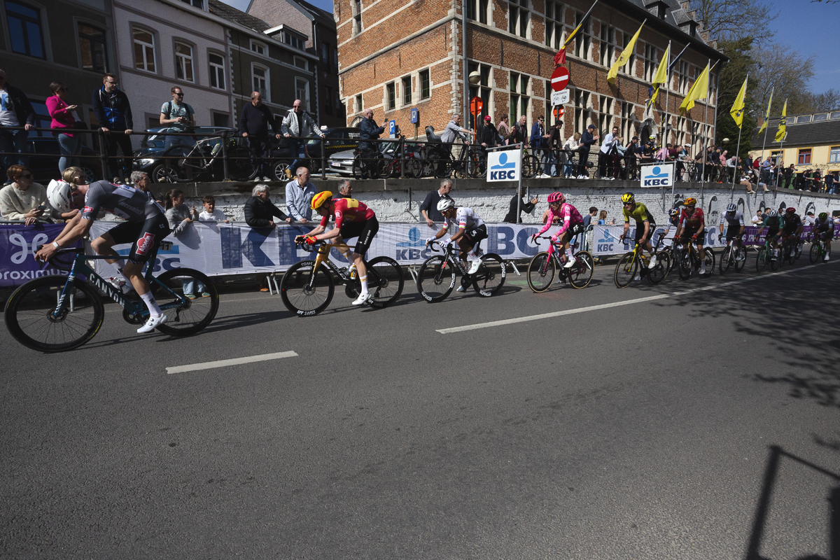De Brabantse Pijl 2025 - A line of riders pass in front of the town hall in Overijse as fans line the streets