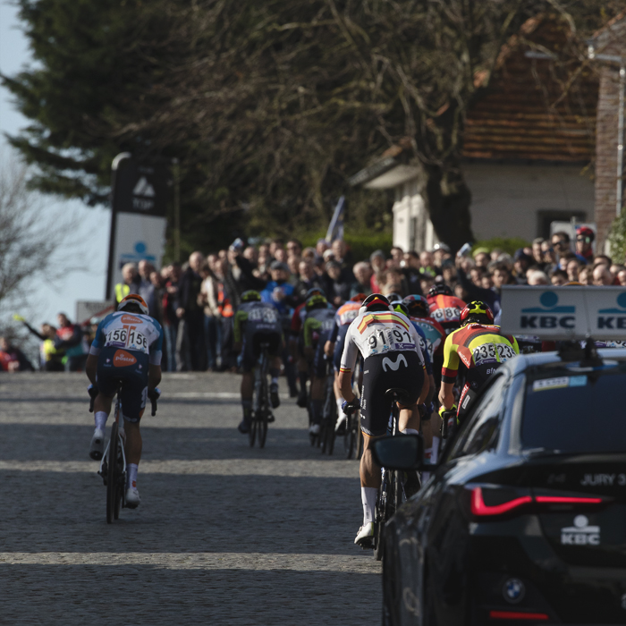 Dwars Door Vlaanderen 2024 - Riders seen from behind followed by a race vehicle up the Nokereberg