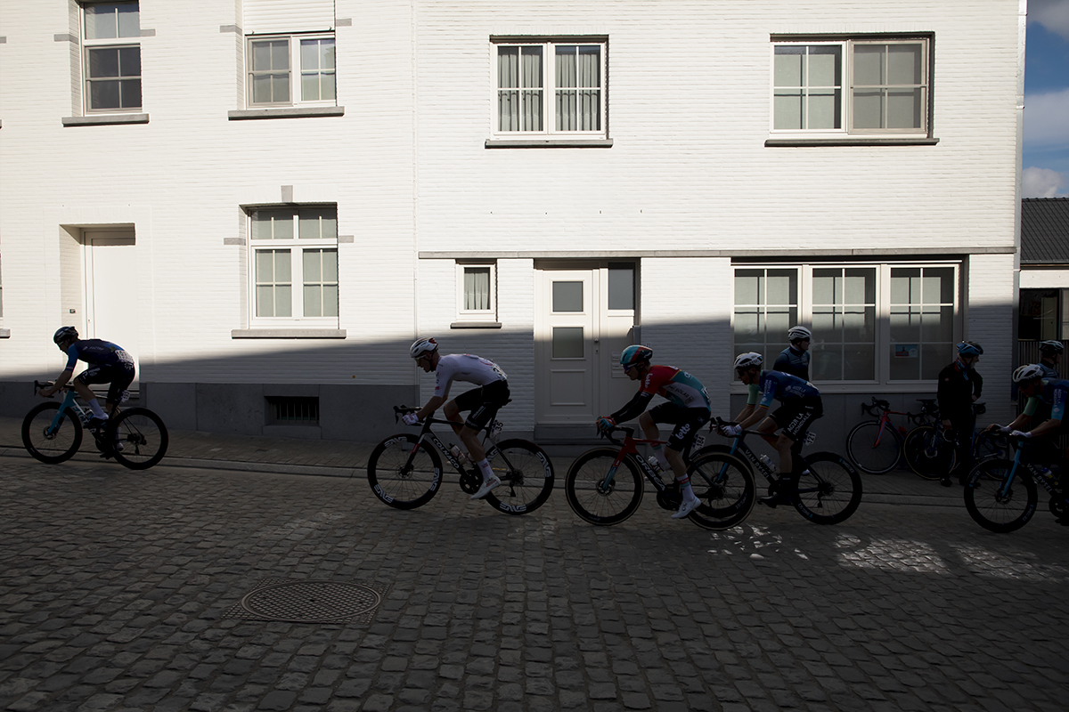 Dwars Door Vlaanderen 2024 - A group of riders in shadow against a bright white washed frontage of a building