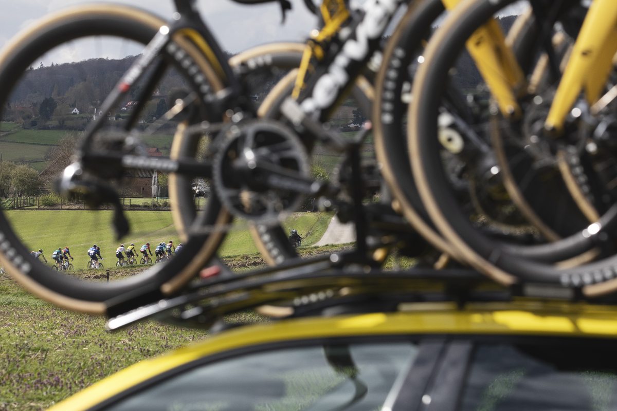 Dwars Door Vlaanderen 2024 - A group of riders on Rue des Chapelles are framed by the wheel of a bike on a team car