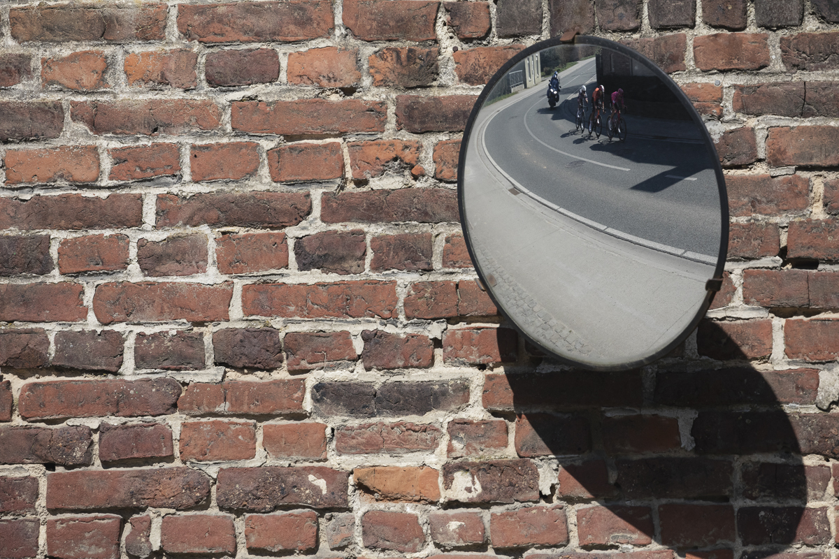Dwars Door Vlaanderen 2025 - Riders reflected in a roadside mirror on a brick wall