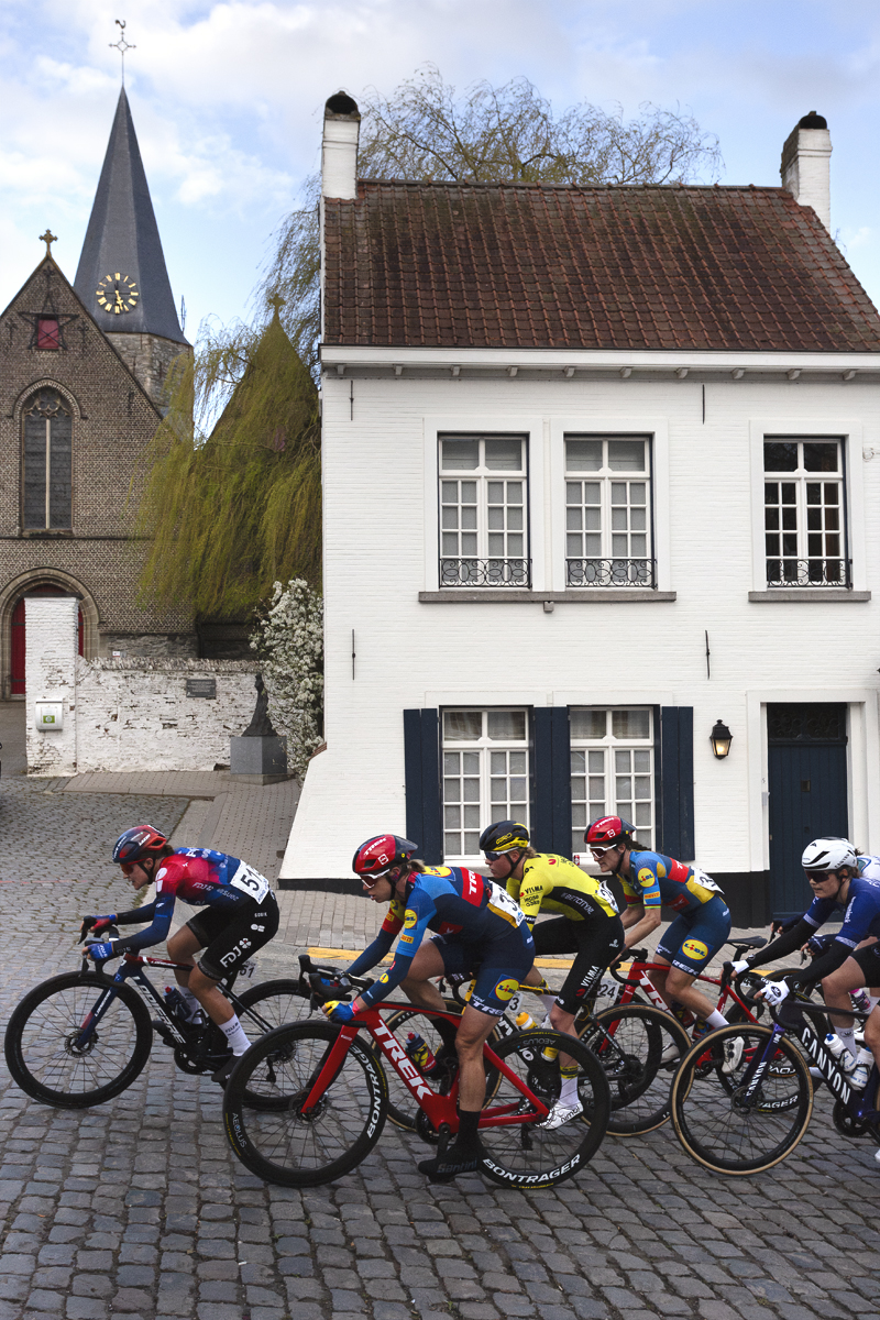 Dwars Door Vlaanderen Vrouwen 2024 - A groups of riders passes a white washed building with shutters in Nokere
