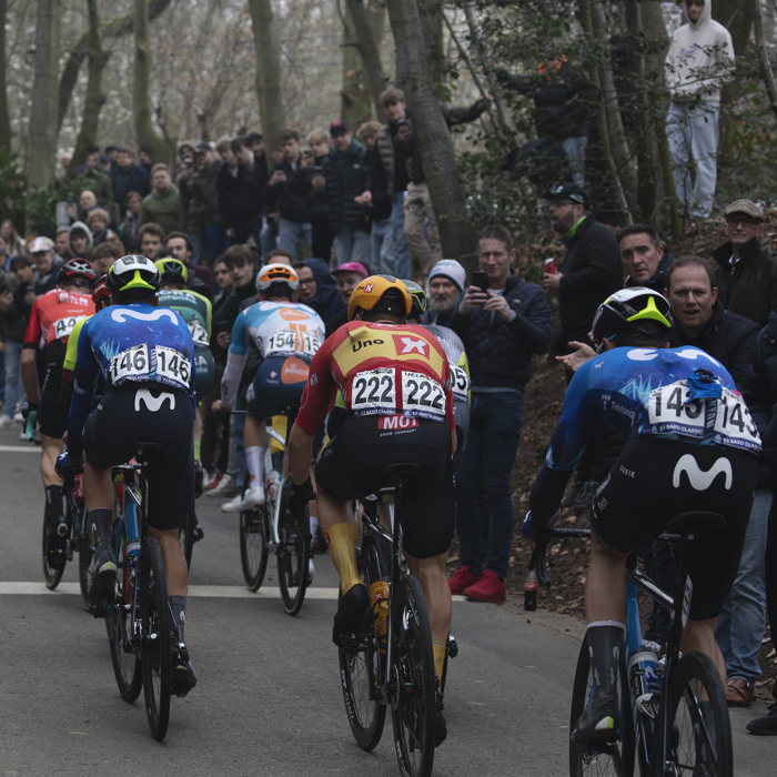 E3 Saxo Classic 2024 - Riders from behind on the climb as fans watch from raised vantage points at the side of the narrow road