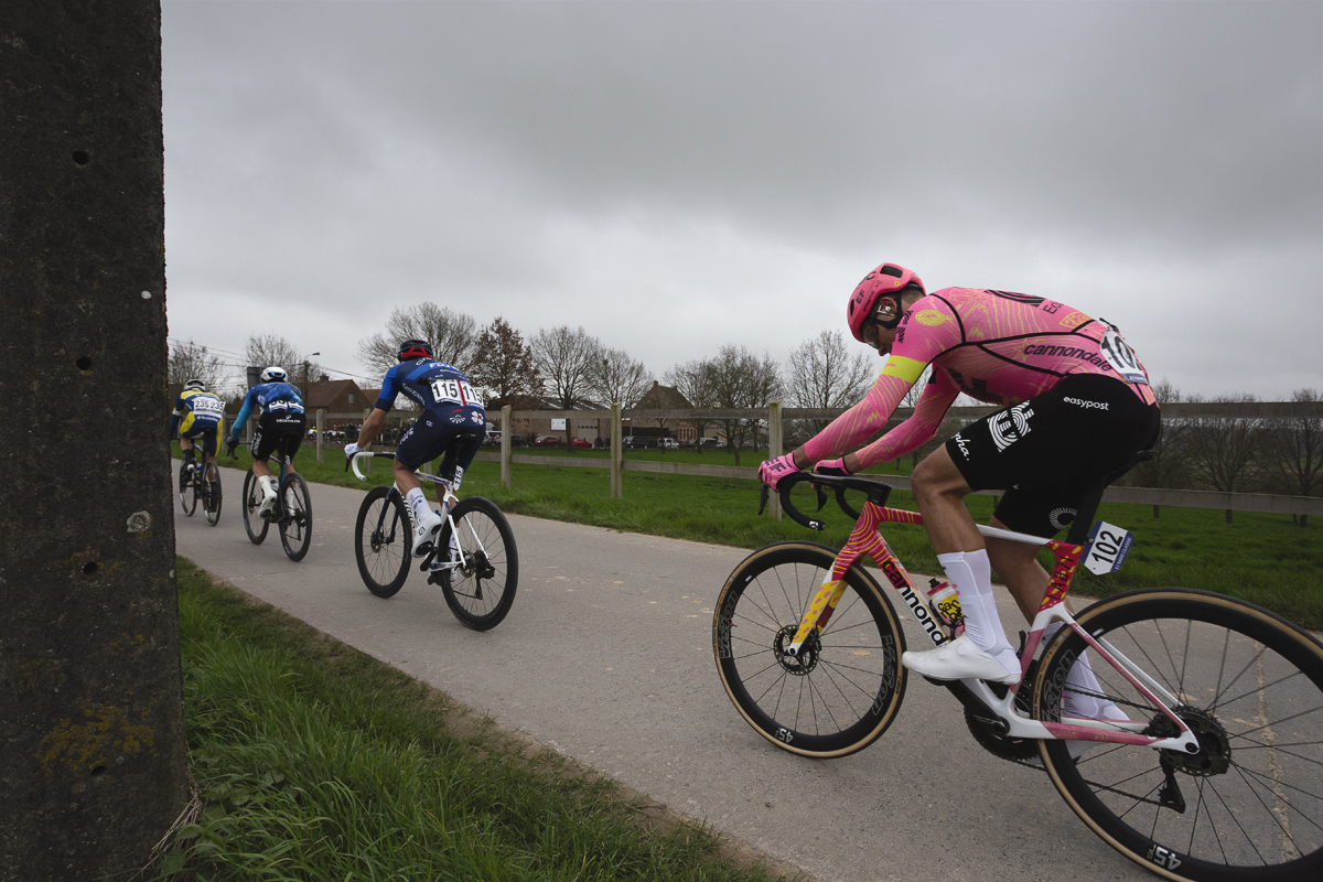 E3 Saxo Classic 2024 - A group of riders pass by a field bordered by a wooden fence