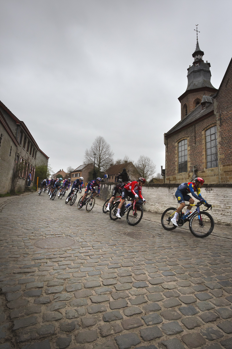 E3 Saxo Classic 2025 - A line of riders on a cobbled road on the way past Sint Denijskerk
