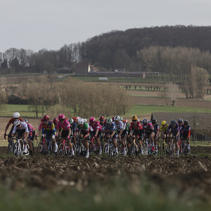 Gent Wevelgem Vrouwen 2025 - Riders seen over a ploughed field with the rise of a distant tree lined hill behind