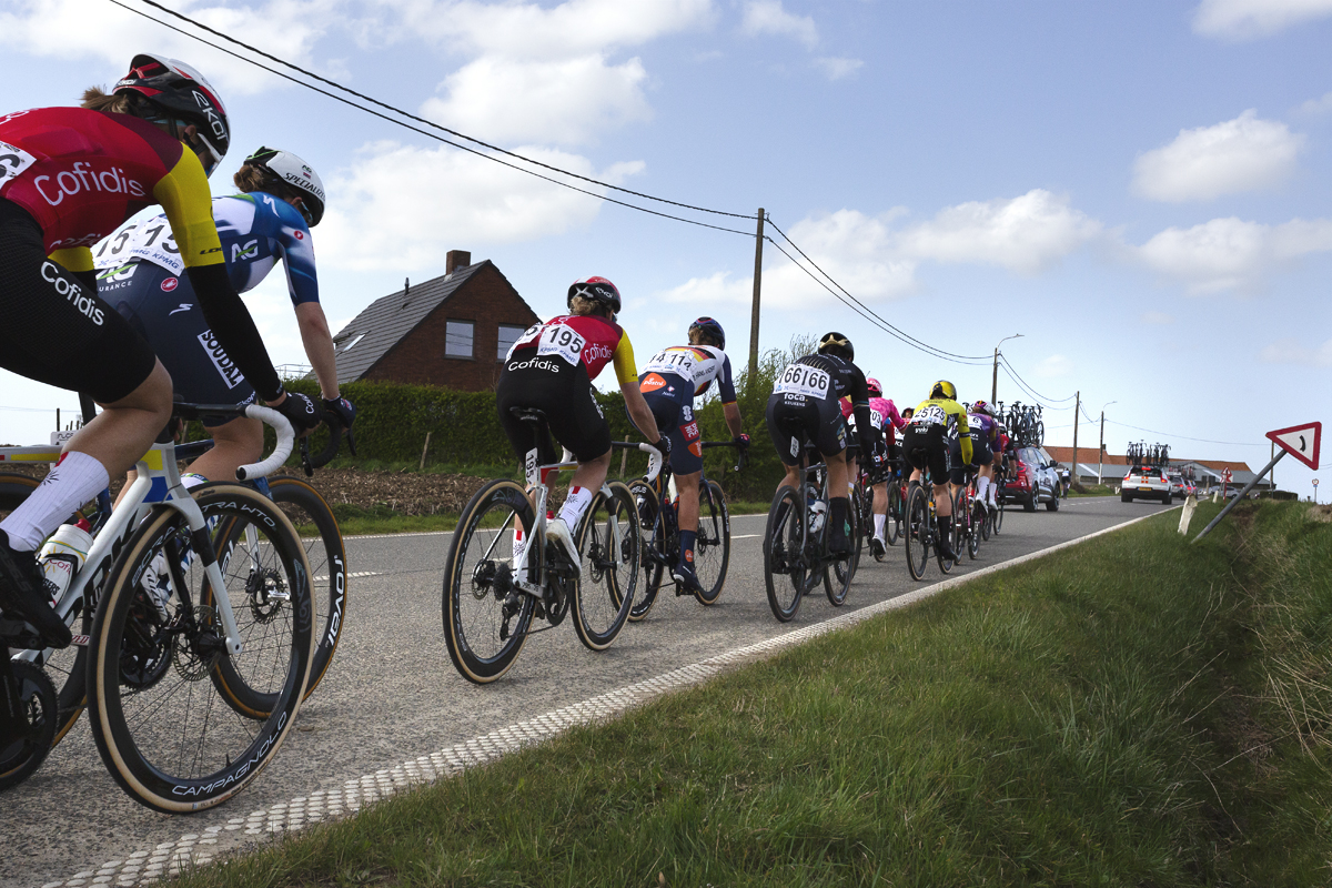 Gent Wevelgem Vrouwen 2025 - Riders from behind with a road sign leaning acutely towards the ditch at one side