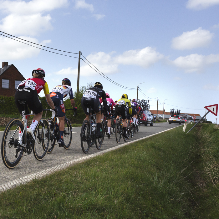 Gent Wevelgem Vrouwen 2025 - Riders from behind with a road sign leaning acutely towards the ditch at one side