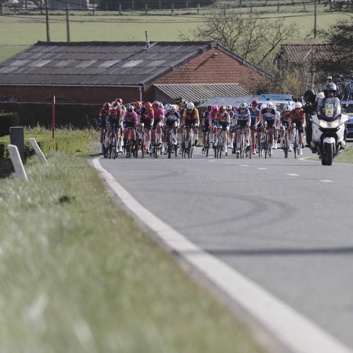 Gent Wevelgem Vrouwen 2025 - Riders approach with old farm buildings behind them in Loker
