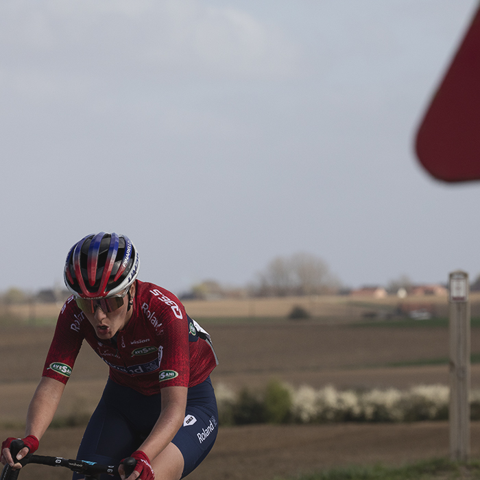 Gent Wevelgem Vrouwen 2025 - Kaja Rysz rides round a bend with the red chevron and red of a road sign matching her kit