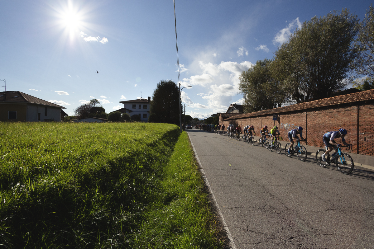 Gran Piemonte 2024 - A line of riders race into Maggiora with a wall to one side and open fields to the other
