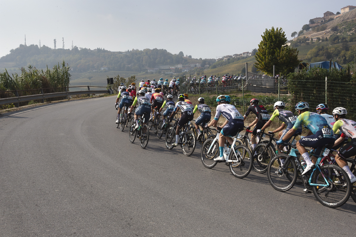 Gran Piemonte 2025 - The peloton rounds a corner with a hill village seen in the distance