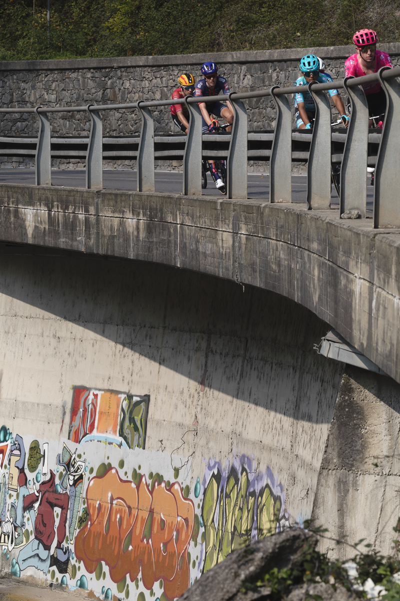 Il Lombardia 2025 - Riders on a raised road with graffiti on the wall beneath them in Ponti