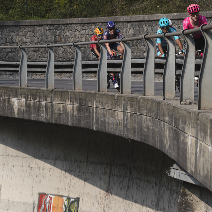 Il Lombardia 2025 - Riders on a raised road with graffiti on the wall beneath them in Ponti