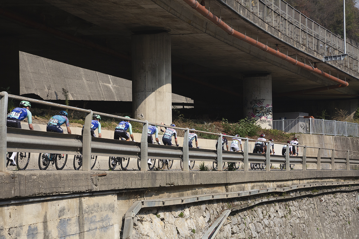 Il Lombardia 2025 - Riders framed by a modern crash barrier pass under a motorway viaduct in Ponti