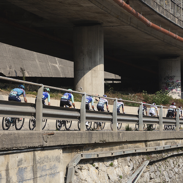 Il Lombardia 2025 - Riders framed by a modern crash barrier pass under a motorway viaduct in Ponti