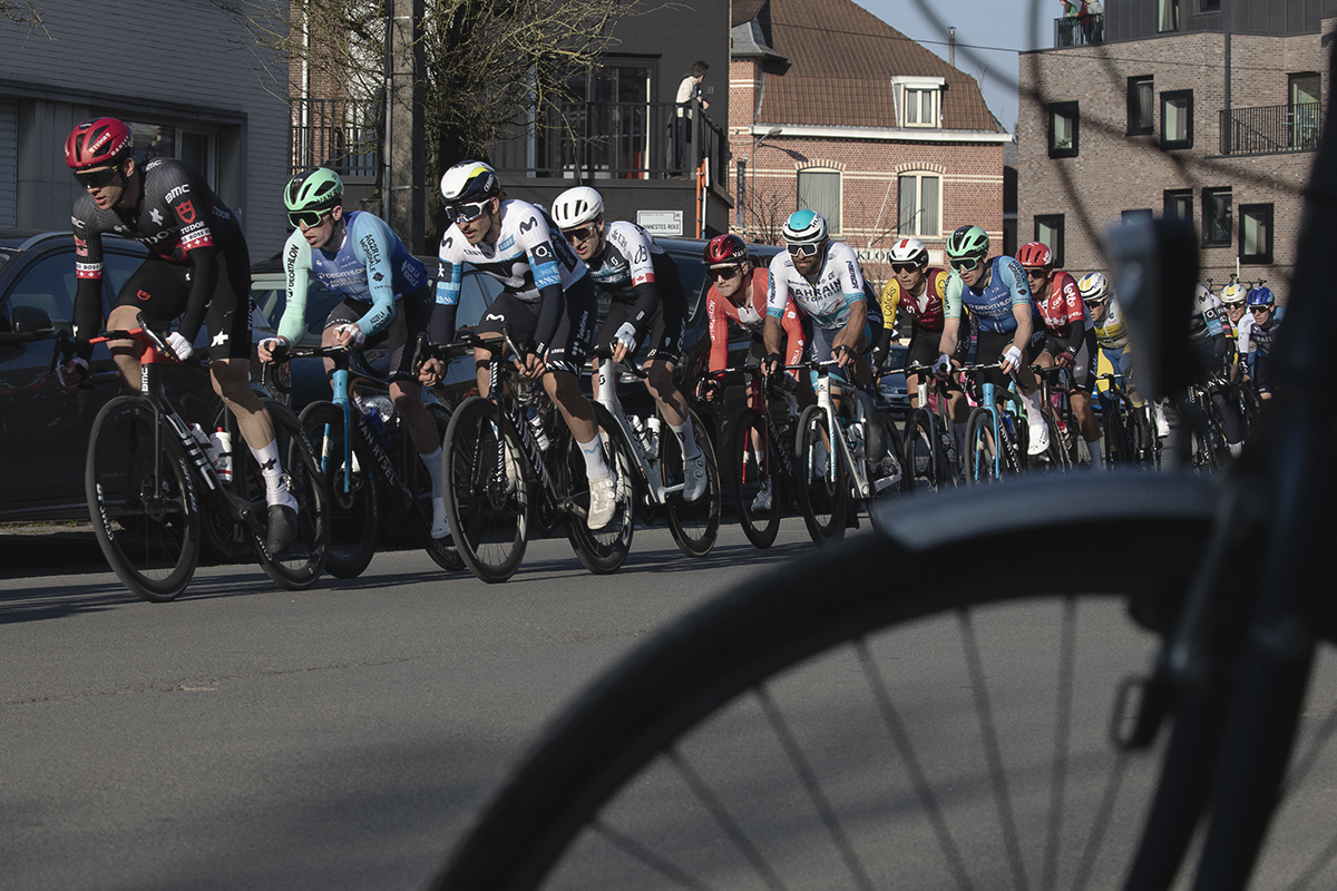 Kuurne Brussel Kuurne 2025 - The peloton races down the street seen through a bike parked in the foreground