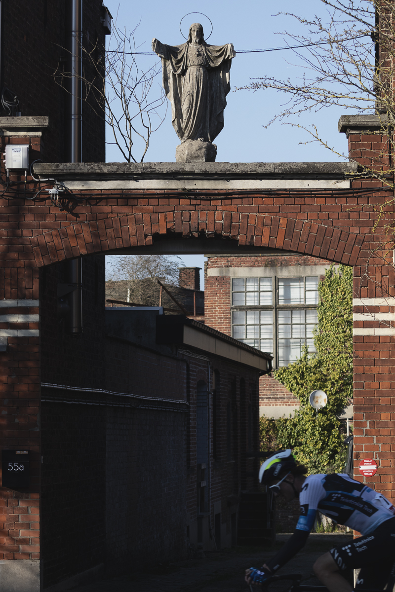 Kuurne Brussel Kuurne 2025 - A rider with his head bowed cycles in front of a brick arch with a statue of Jesus adorning it