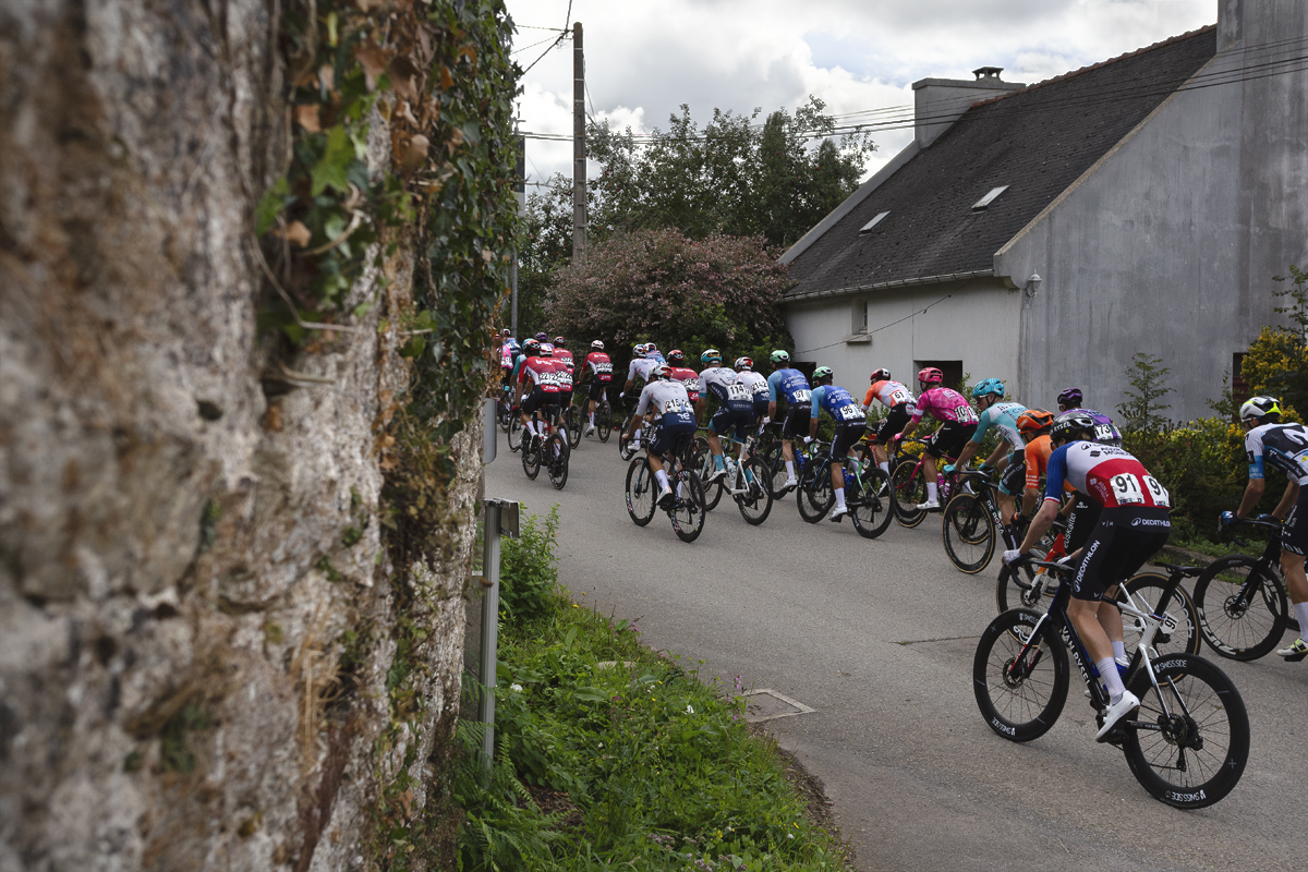 Bretagne Classic - Ouest-France 2025 - The peloton moves away down the road framed by a stone wall and small cottage