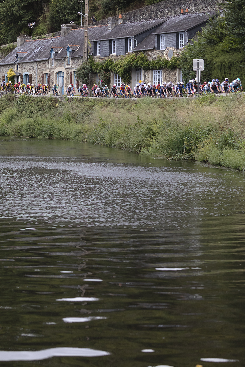 Bretagne Classic - Ouest-France 2025 - The peloton viewed over the water passing in front of a row of stone cottages on the Quai Jean Guivarc'h
