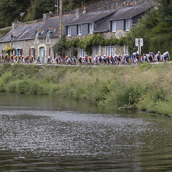 Bretagne Classic - Ouest-France 2025 - The peloton viewed over the water passing in front of a row of stone cottages on the Quai Jean Guivarc'h