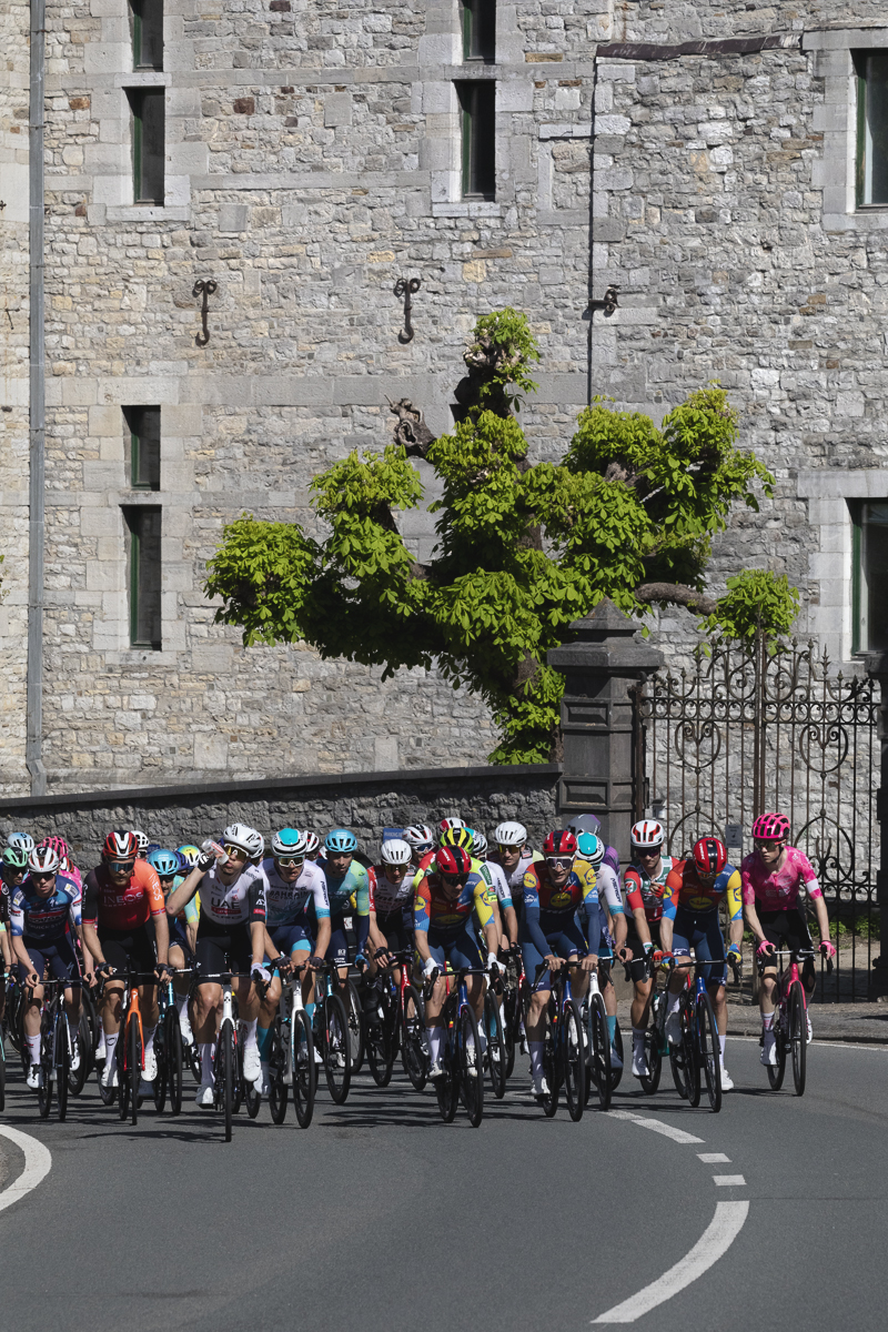 Liege Bastogne Liege 2025 - A stone wall of Château de Harzé looms in the background as the riders pass its iron gates