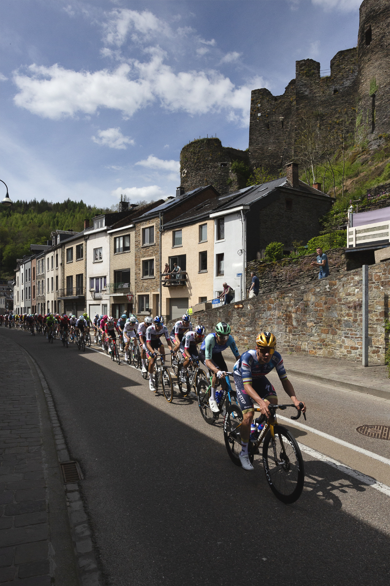 Liege Bastogne Liege 2025 - The peloton passes a row of houses set into the rock face beneath the castle at La Roché-en-Ardenne