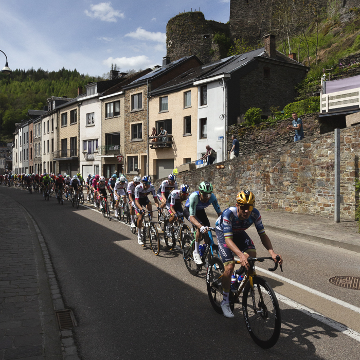Liege Bastogne Liege 2025 - The peloton passes a row of houses set into the rock face beneath the castle at La Roché-en-Ardenne
