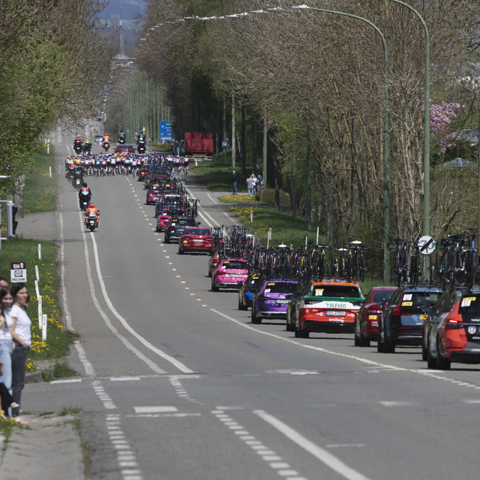 Liège Bastogne Liège Femmes 2025 - The race convoy lines out down a long straight road with riders in the distance in Foy