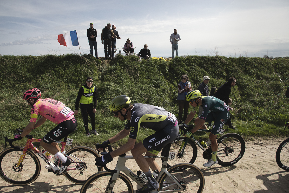 Paris Roubaix 2024 - Fans fly a Tricolour as they stand on a grassy bank to watch the race on the Quiévy à Saint-Python sector