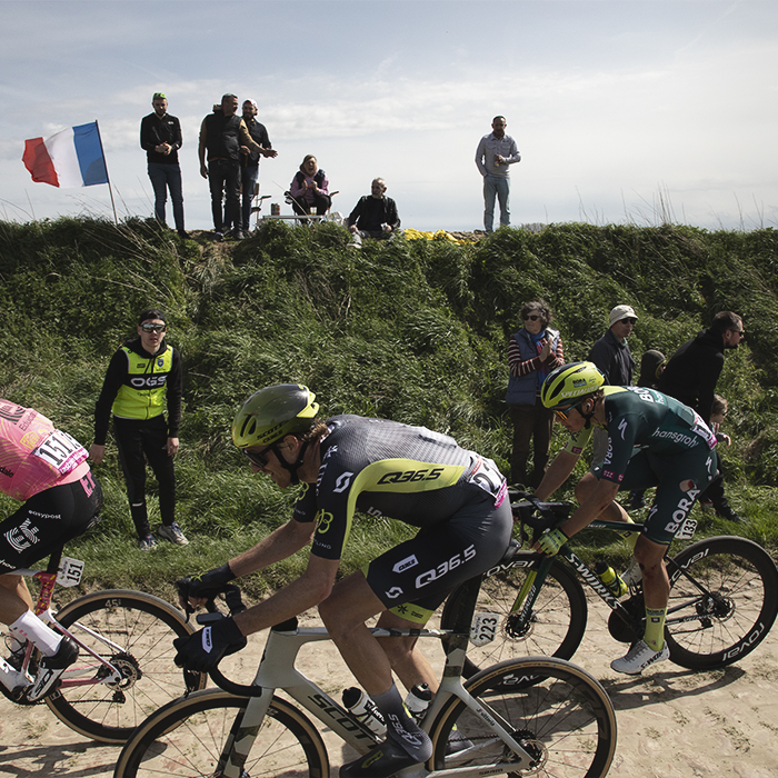 Paris Roubaix 2024 - Fans fly a Tricolour as they stand on a grassy bank to watch the race on the Quiévy à Saint-Python sector