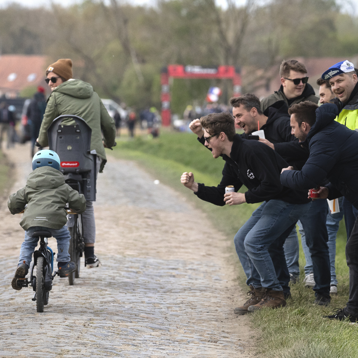 Paris Roubaix 2025 - A young fan and their mother are cheered on by a group of fans as they ride on the cobbles