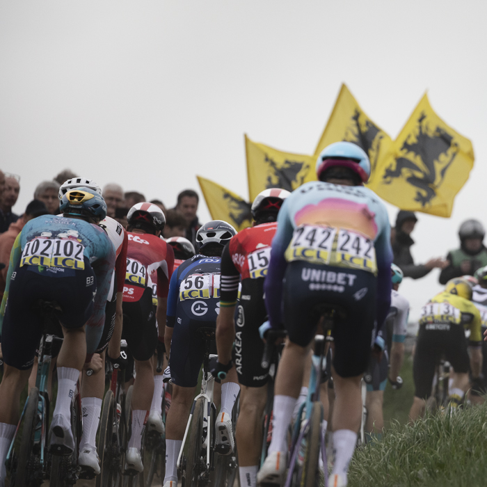 Paris Roubaix 2025 - A rear view riders with the flags of Flanders flying on Quérénaing à Maing