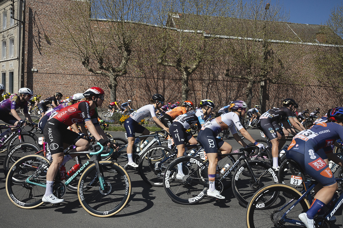 Paris Roubaix Femmes 2025 - The peloton rounds a bend with trees just coming into bud in the background