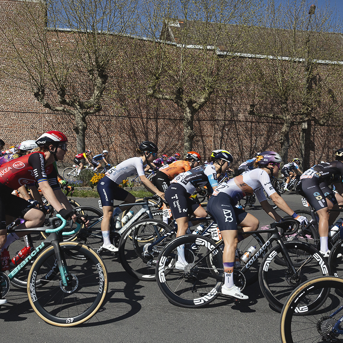 Paris Roubaix Femmes 2025 - The peloton rounds a bend with trees just coming into bud in the background