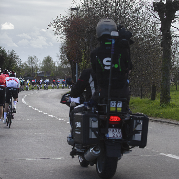 Ronde van Vlaanderen 2024 - The curve of the riders as they take a sweeping bend can be seen as the race is followed by a TV Moto