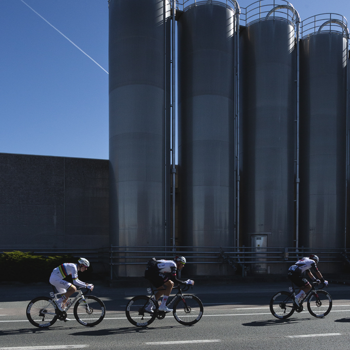 Ronde van Vlaanderen 2025 - A group of elite riders race in front of a line of industrial silos